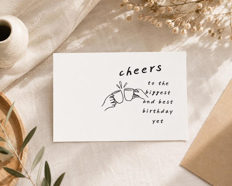 May include: A white rectangular greeting card featuring a black line drawing of two hands clinking mugs together. The text on the card reads: cheers to the biggest and best birthday yet. The design is centered on the front of the card.