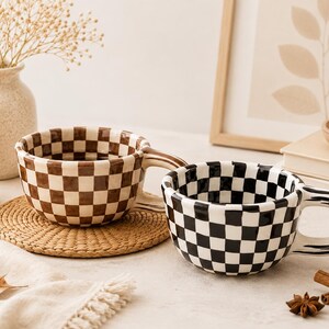May include: Two ceramic mugs with rounded shapes and wide handles. One mug features a brown and white checkered pattern, while the other displays a black and white checkered pattern. Both mugs have a glossy finish and a consistent grid design.
