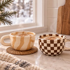 May include: Two ceramic mugs with rounded handles, one featuring a beige and cream abstract wavy pattern and the other a brown and cream checkerboard design. Both mugs are filled with a warm beverage topped with foam and a dusting of cinnamon.