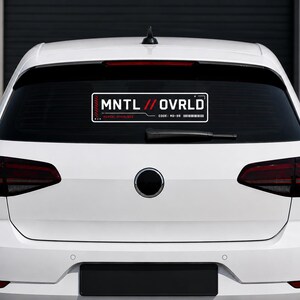 Könnte beinhalten: Ein weißes Auto mit einer schwarzen Heckscheibe, auf der ein rechteckiger Aufkleber angebracht ist. Der Aufkleber trägt den weißen Text "MNTL // OVRLD" mit roten und schwarzen Akzenten. Darunter befindet sich der Code "MD-59" und ein Barcode.