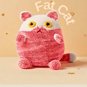May include: A handmade, plush cat toy in pink and white, with yellow eyes and a grey tail. The text "Fat Cat" is above the cat. The cat is sitting on a light yellow surface, with gold star decorations.