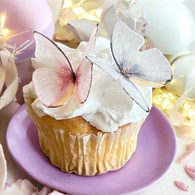 May include: A cupcake with white frosting and two butterfly decorations. The butterflies have delicate, translucent wings with pink and gray hues. The cupcake sits on a small, round, lavender plate.