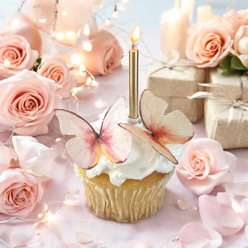 May include: A cupcake with white frosting, topped with two butterfly decorations and a lit gold candle. The cupcake is surrounded by pink roses, gift boxes, and fairy lights, creating a celebratory atmosphere.