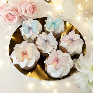 May include: A golden plate holds six cupcakes topped with white frosting and delicate butterfly decorations. The butterflies feature pastel colors like pink, blue, and peach. Pink roses and fairy lights are in the background.