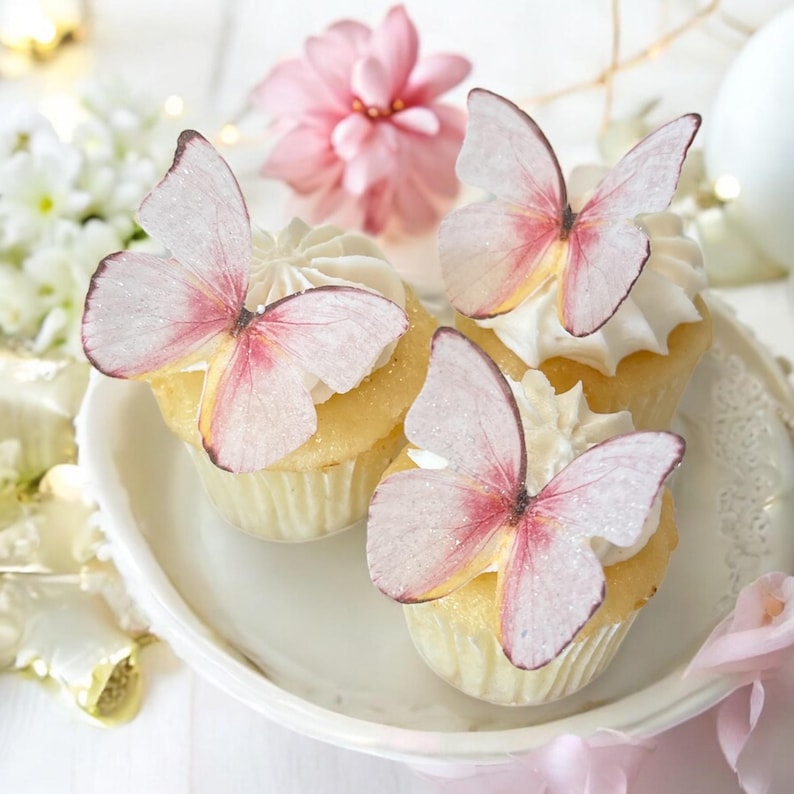 May include: Three cupcakes with white frosting and butterfly decorations. The butterfly wings are light pink with darker pink accents and a touch of glitter. The cupcakes sit on a white, scalloped-edge plate.