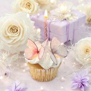 May include: A cupcake with white frosting, decorated with two butterfly toppers and a lit pink candle. The cupcake is surrounded by white roses, a wrapped gift, and fairy lights, creating a celebratory theme.