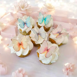 May include: Six cupcakes with white frosting, each topped with a delicate, colorful butterfly decoration. The butterflies feature shades of blue, pink, orange, and purple. The cupcakes sit on a pink surface, with soft lighting and floral accents.
