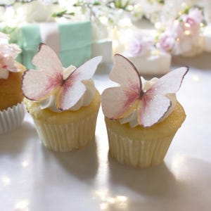 May include: Two cupcakes with white frosting, topped with delicate butterfly decorations. The butterflies have pink and white wings with glitter accents. The cupcakes sit on a white surface, with blurred decorations in the background.