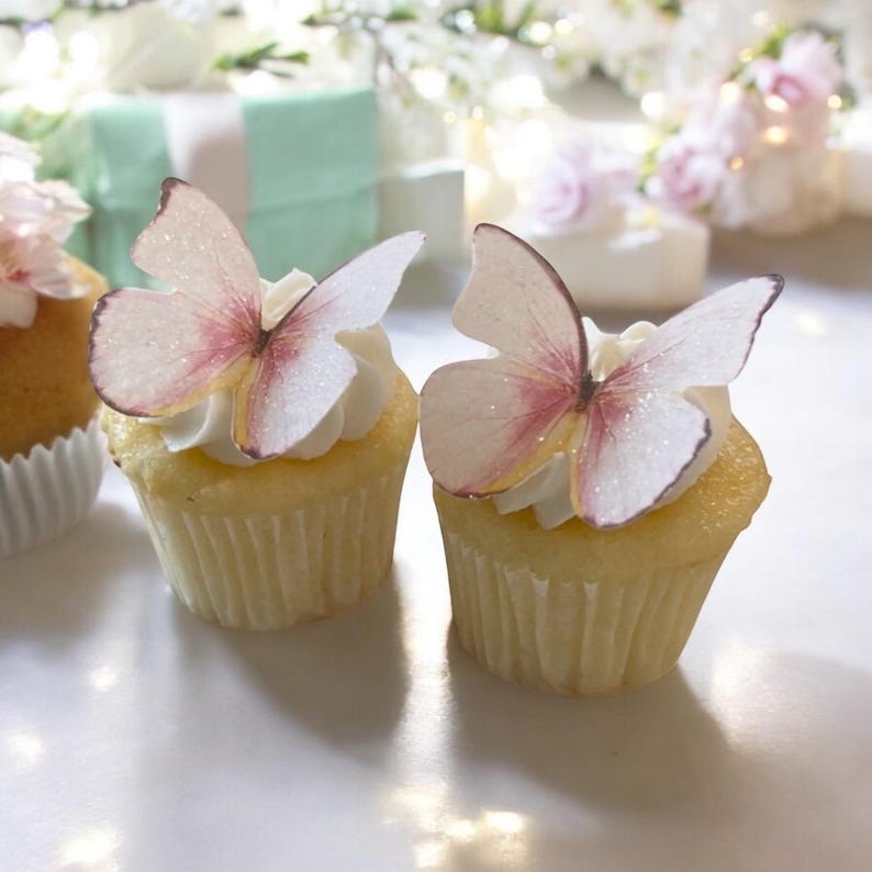 May include: Two cupcakes with white frosting, topped with delicate butterfly decorations. The butterflies have pink and white wings with glitter accents. The cupcakes sit on a white surface, with blurred decorations in the background.