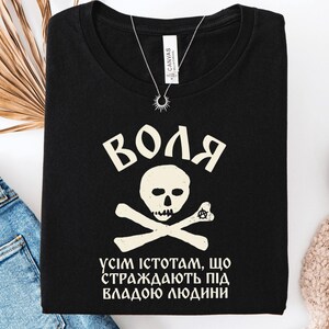 May include: A black t-shirt featuring a cream-colored graphic of a skull and crossbones. Above the skull is the Cyrillic text "ВОЛЯ". Below the skull is the Cyrillic text "УСІМ ІСТОТАМ, ЩО СТРАЖДАЮТЬ ПІД ВЛАДОЮ ЛЮДИНИ". The shirt has a crew neckline and a white label.