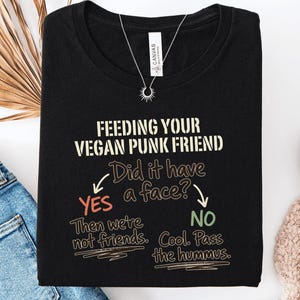 May include: A black t-shirt featuring the text FEEDING YOUR VEGAN PUNK FRIEND. Below, a flowchart asks Did it have a face? with arrows pointing to YES, then we're not friends, and NO, Cool. Pass the hummus. The design uses white, red, and green lettering.