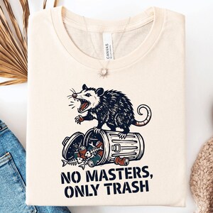 May include: A cream-colored t-shirt featuring a graphic of a snarling opossum standing on a tipped-over metal trash can filled with debris. Below the illustration, the text reads NO MASTERS, ONLY TRASH in bold, dark blue, capital letters.