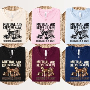 May include: Six folded t-shirts in cream, pink, light blue, black, maroon, and navy blue. Each shirt features a graphic of a dog pulling a cart with supplies and rats, with the text "MUTUAL AID KEEPS US ALIVE" and "HOUSING IS A RIGHT" printed in black or tan.