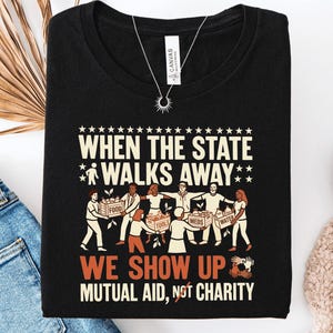 May include: A black t-shirt featuring a graphic print with the text "WHEN THE STATE WALKS AWAY" and "WE SHOW UP MUTUAL AID, NOT CHARITY". The illustration depicts a group of people holding crates labeled "FOOD", "TOOLS", "MEDS", and "WATER".