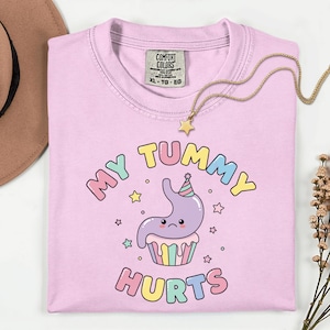 May include: A light pink t-shirt with the text "MY TUMMY HURTS" in colorful bubble letters. The shirt features a cartoon stomach character wearing a party hat and sitting on a cupcake. A gold necklace with a star pendant is draped across the shirt.