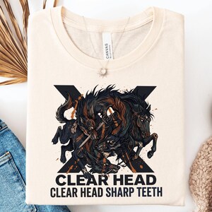 May include: A cream-colored t-shirt featuring a graphic of a black wolf and a black horse intertwined with a sword. The design includes a large black X behind the animals and the text "CLEAR HEAD" and "CLEAR HEAD SHARP TEETH" in black.