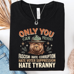 May include: A black t-shirt featuring a graphic of a brown bear wearing a green ranger hat. The text reads: ONLY YOU CAN PREVENT FASCISM HATE CORRUPTION HATE VOTER SUPPRESSION HATE TYRANNY. The design includes small star and pine branch accents.