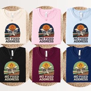 May include: Six crew neck t-shirts in cream, pink, light blue, black, maroon, and navy blue. Each shirt features a graphic of two birds perched on a wire against a sunset with an anarchy symbol and the text NO FIXED ADDRESS printed below.