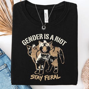May include: A black t-shirt featuring a graphic of a cat with a mohawk and spiked collar. The cat is filled with a newspaper-style print. Above the cat is the text "GENDER IS A RIOT" and below is the text "STAY FERAL" in a tan, distressed font.