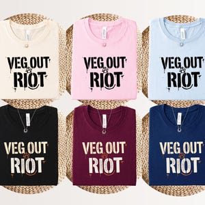 May include: Six crew neck t-shirts in cream, light pink, light blue, black, maroon, and navy blue. Each shirt features the black or white text "VEG OUT RIOT" with a circular anarchy symbol replacing the letter O, designed in a distressed graffiti style.
