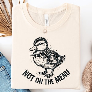 May include: A cream-colored t-shirt featuring a black graphic illustration of a duckling wearing a spiked collar. Below the duckling, the text reads "NOT ON THE MENU" in a bold, distressed black font. The shirt has a crew neckline and short sleeves.