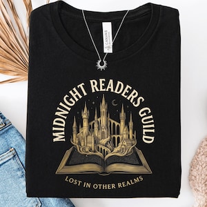 May include: A black t-shirt featuring a graphic of an open book with a castle rising from its pages. The text reads "MIDNIGHT READERS GUILD" in an arch above the castle and "LOST IN OTHER REALMS" in a straight line below the open book.