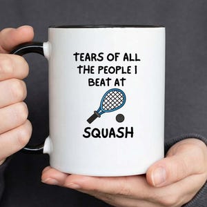 Könnte beinhalten: Weißer Keramikbecher mit schwarzem Innenraum und Henkel. Der Becher zeigt den Text "TEARS OF ALL THE PEOPLE I BEAT AT SQUASH" zusammen mit einer blauen Squashschläger- und Ballgrafik. Der Becher wird von einer Person gehalten.