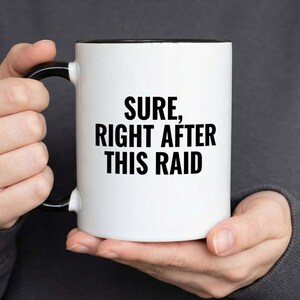 Puede incluir: Taza de cerámica blanca con interior y asa negros. La taza presenta el texto "SURE, RIGHT AFTER THIS RAID" en negrita negra. La taza es sostenida por una persona que lleva una sudadera gris oscura.