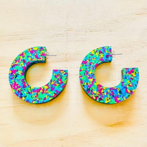 May include: A pair of colourful, C-shaped earrings. The earrings have a confetti-like pattern with blue, green, yellow, and pink hues. The earrings have a black border and a silver post for pierced ears. The earrings are on a light wood surface.