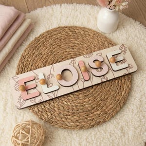 May include: A wooden name puzzle spelling "Eloise" in pink and white letters with wooden pegs. The puzzle is on a floral patterned background with a light beige colour. The puzzle is placed on a woven mat.