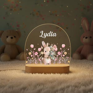 May include: A decorative night light featuring a bunny holding flowers, with the name "Lydia" in white script. The light has a wooden base and a clear, arched top with floral and butterfly accents. Two stuffed animals are in the background.