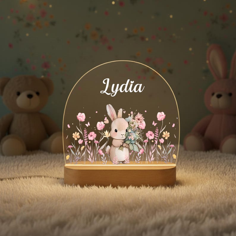May include: A decorative night light featuring a bunny holding flowers, with the name "Lydia" in white script. The light has a wooden base and a clear, arched top with floral and butterfly accents. Two stuffed animals are in the background.