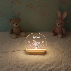 May include: A personalized night light with the name "Lydia" displayed on a clear, arched panel. The panel features a bunny holding flowers amidst floral illustrations. The light sits on a wooden base, with a teddy bear and a pink bunny plush toy in the background.