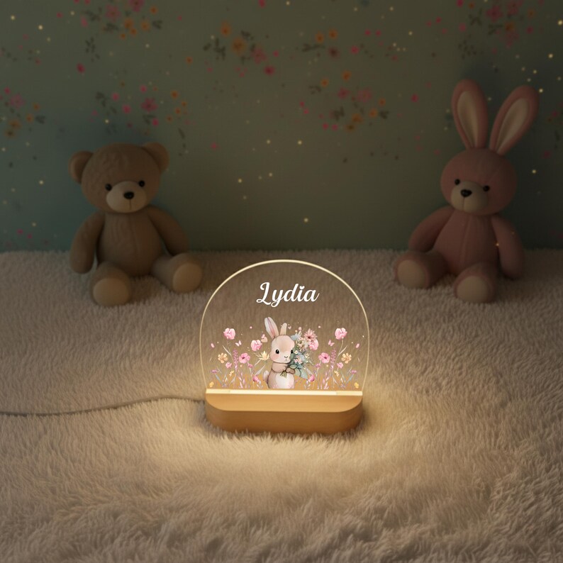 May include: A personalized night light with the name "Lydia" displayed on a clear, arched panel. The panel features a bunny holding flowers amidst floral illustrations. The light sits on a wooden base, with a teddy bear and a pink bunny plush toy in the background.