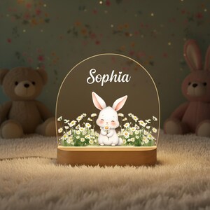 May include: A night light featuring a white bunny holding a daisy, surrounded by daisies, with the name "Sophia" above. The light sits on a wooden base and is surrounded by stuffed animals.
