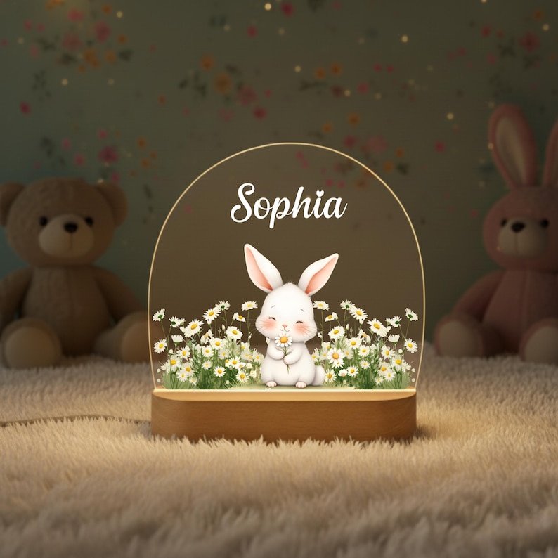 May include: A night light featuring a white bunny holding a daisy, surrounded by daisies, with the name "Sophia" above. The light sits on a wooden base and is surrounded by stuffed animals.
