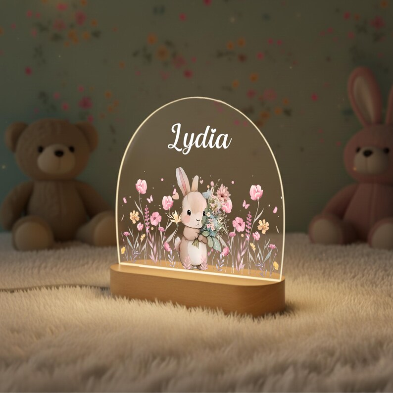 May include: A personalized night light with the name "Lydia" in white script. The acrylic panel features a watercolor illustration of a bunny holding flowers, surrounded by pink and yellow flowers. The base is made of light-colored wood.