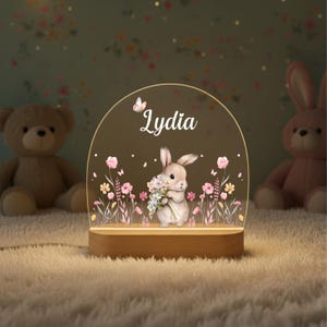 May include: A personalized night light featuring a bunny holding flowers, with the name "Lydia" in script. The design includes pink and yellow flowers, butterflies, and a wooden base. A teddy bear and a pink bunny are in the background.