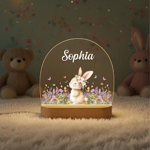 May include: A decorative night light featuring a smiling bunny with a floral crown, surrounded by colorful flowers and butterflies. The name "Sophia" is displayed above the bunny. The light sits on a wooden base.