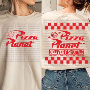 May include: Cream-colored sweatshirt with a red graphic of "Pizza Planet" and a planet with a pizza slice. The back features a red and white checkered pattern and the words "Delivery Shuttle."