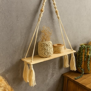 Puede incluir: Estante colgante de madera con detalles de macramé. El estante está suspendido por una cuerda trenzada y presenta un acabado de madera natural. Contiene un jarrón texturizado, un pequeño recipiente de madera y plantas secas. Borlas cuelgan de la parte inferior del estante.