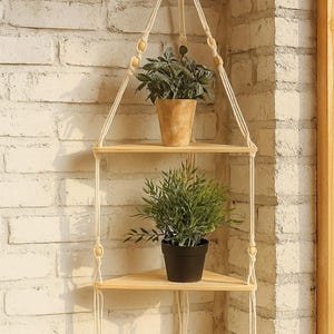 May include: A two-tiered, triangular hanging shelf made of light wood, suspended by off-white macrame rope. The shelf holds two potted plants, one in a terracotta pot and the other in a black pot, against a white brick wall.