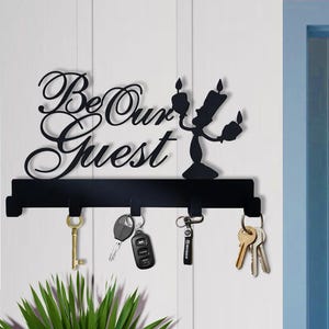 Puede incluir: Llavero de metal negro con las palabras "Be Our Guest" en escritura elegante, con un diseño de candelabro. Varias llaves cuelgan de los ganchos. El llavero está montado en una pared blanca, con una puerta azul al fondo.