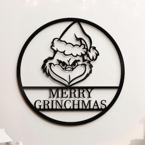 Pode incluir: Decoração de parede em metal preto com o tema Grinch. O design apresenta o rosto do Grinch e seu gorro de Papai Noel dentro de uma moldura circular, com as palavras "MERRY GRINCHMAS" abaixo. Uma decoração festiva de Natal.