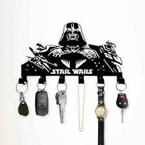 May include: Black metal key holder featuring a Star Wars design with Darth Vader. The key holder has multiple hooks with keys, a watch, and a necklace hanging from them. The key holder is mounted on a white wall.