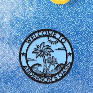 以下が含まれることがあります： 青い背景に、黄色の浮き輪と、ヤシの木と波のデザインで「WELCOME TO ANDERSON'S OASIS EST.2025」と書かれた円形の看板があります。看板は黒で、背景は水色です。
