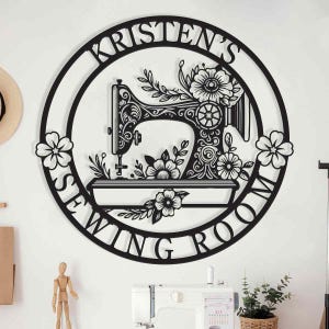 May include: Black metal wall art with the text "Kristen's Sewing Room" encircling a detailed sewing machine design. Floral accents are incorporated throughout the design, adding a decorative touch to the piece. The artwork is mounted on a white wall.
