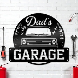 May include: Black metal sign with the words "Dad's Garage" in a stylized font. The sign features a truck silhouette and a forest scene, mounted on a white brick wall. Tools and a red gas can are visible.