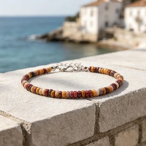 Pode incluir: Uma pulseira de contas composta por pequenas contas circulares em forma de disco plano numa paleta de cores repetitivas de vermelho profundo, amarelo mostarda e creme suave. A pulseira é finalizada com um fecho lagosta de metal prateado.