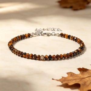 Könnte beinhalten: Ein Armband aus kleinen, facettierten, runden Tigerauge-Edelsteinen in Braun-, Gold- und Schwarztönen. Das Armband verfügt über eine silberfarbene Metall-Verlängerungskette und einen Karabinerverschluss, die eine individuelle Anpassung der Länge ermöglichen.
